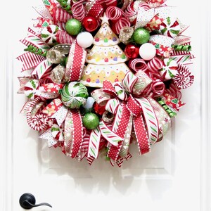 Puede incluir: Una corona navide&ntilde;a festiva con cintas rojas, blancas y verdes, adornos y un &aacute;rbol de jengibre. La corona est&aacute; adornada con detalles de bastones de caramelo y varios elementos decorativos, creando una exhibici&oacute;n navide&ntilde;a alegre.