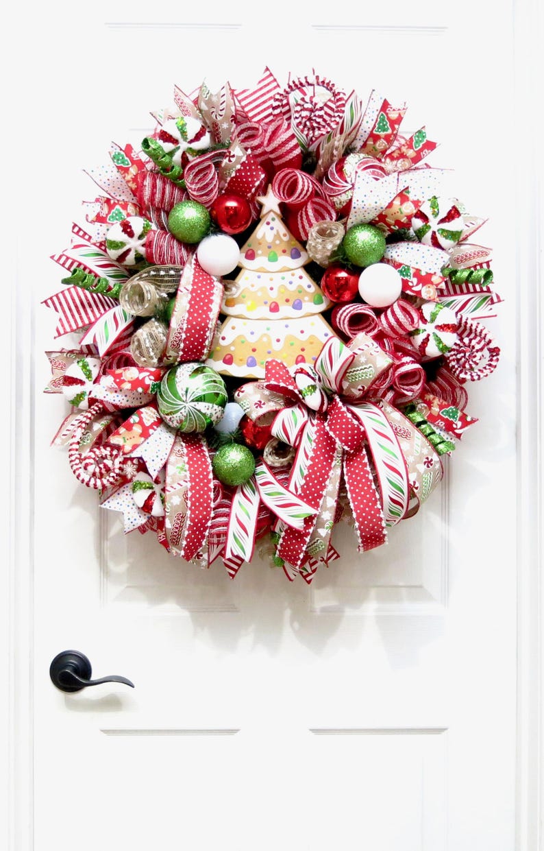 Puede incluir: Una corona navide&ntilde;a festiva con cintas rojas, blancas y verdes, adornos y un &aacute;rbol de jengibre. La corona est&aacute; adornada con detalles de bastones de caramelo y varios elementos decorativos, creando una exhibici&oacute;n navide&ntilde;a alegre.