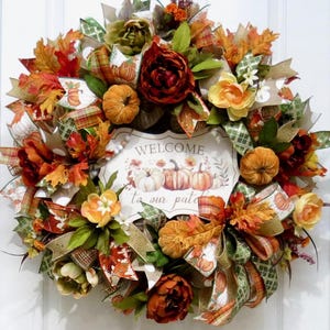 May include: A fall-themed wreath with orange, red, and green leaves, pumpkins, and flowers. The wreath has a sign that reads "WELCOME to our patch". Ribbons and decorative elements complete the festive design.