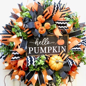 Puede incluir: Una corona oto&ntilde;al festiva con cintas naranjas, negras y blancas, calabazas artificiales y vegetaci&oacute;n. Un letrero negro en el centro dice "hello PUMPKIN" con una ilustraci&oacute;n de calabaza. Un art&iacute;culo decorativo para la temporada de oto&ntilde;o.