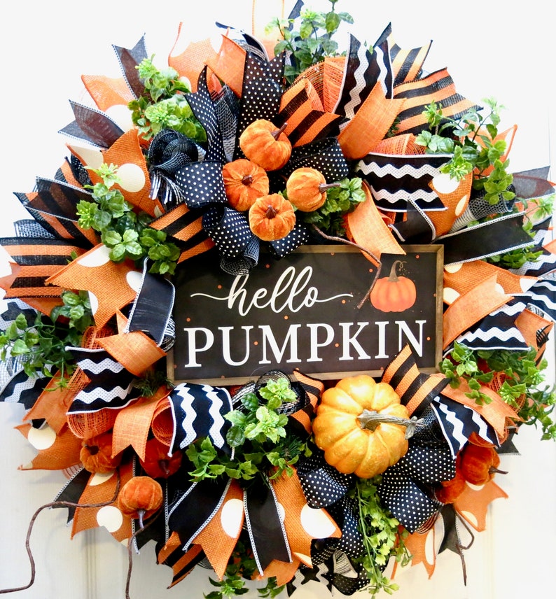 Puede incluir: Una corona oto&ntilde;al festiva con cintas naranjas, negras y blancas, calabazas artificiales y vegetaci&oacute;n. Un letrero negro en el centro dice "hello PUMPKIN" con una ilustraci&oacute;n de calabaza. Un art&iacute;culo decorativo para la temporada de oto&ntilde;o.