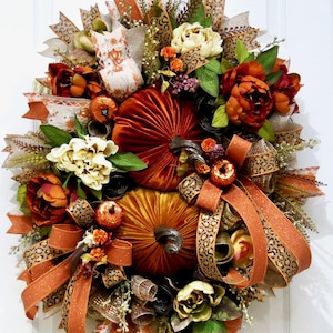 May include: A decorative wreath featuring two large velvet pumpkins in rust and gold, surrounded by cream and dark orange peonies, small orange gourds, and various ribbons with leopard print, glittery orange, and neutral patterns on a wire frame.