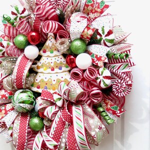 Puede incluir: Una corona navide&ntilde;a festiva con cintas rojas, blancas y verdes, detalles de bastones de caramelo y adornos decorativos. La corona presenta un centro de &aacute;rbol de pan de jengibre para una exhibici&oacute;n navide&ntilde;a.