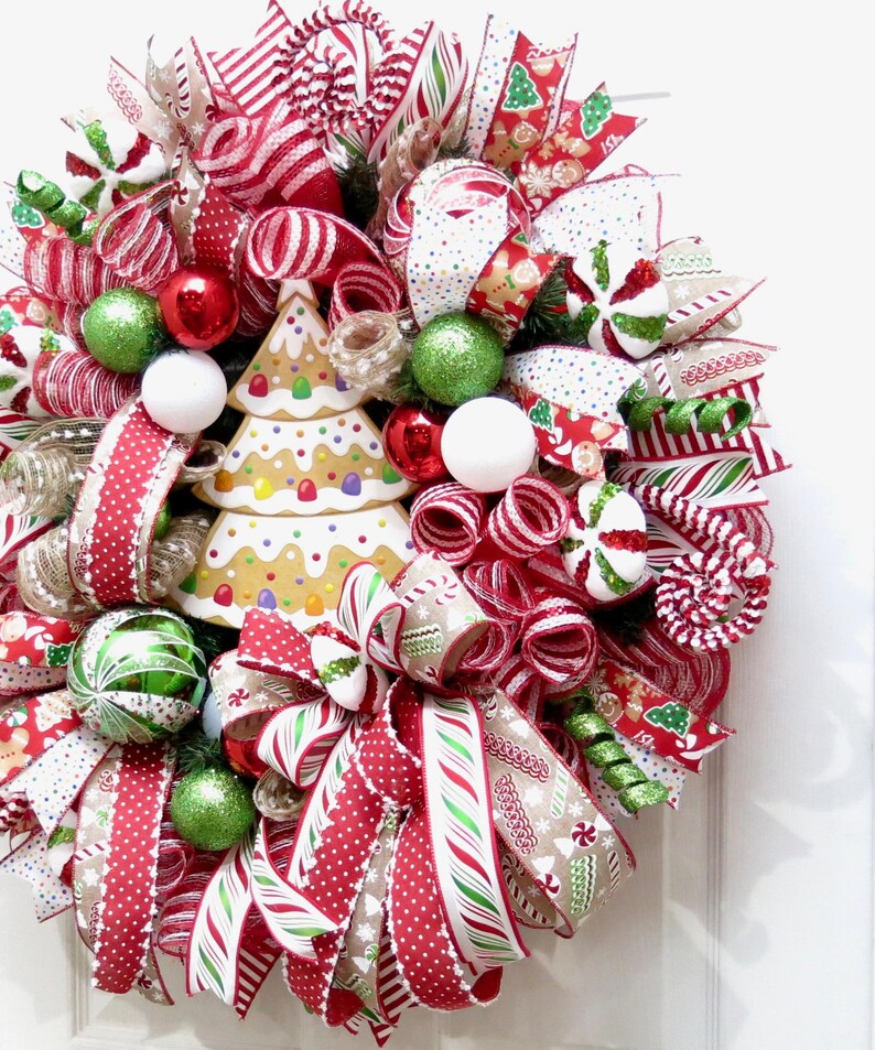 Puede incluir: Una corona navide&ntilde;a festiva con cintas rojas, blancas y verdes, detalles de bastones de caramelo y adornos decorativos. La corona presenta un centro de &aacute;rbol de pan de jengibre para una exhibici&oacute;n navide&ntilde;a.