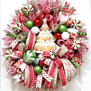 Puede incluir: Una festiva corona navide&ntilde;a con un centro de mesa de &aacute;rbol de pan de jengibre. La corona presenta cintas a rayas rojas y blancas, adornos verdes y rojos y detalles de bastones de caramelo. Un art&iacute;culo decorativo navide&ntilde;o.