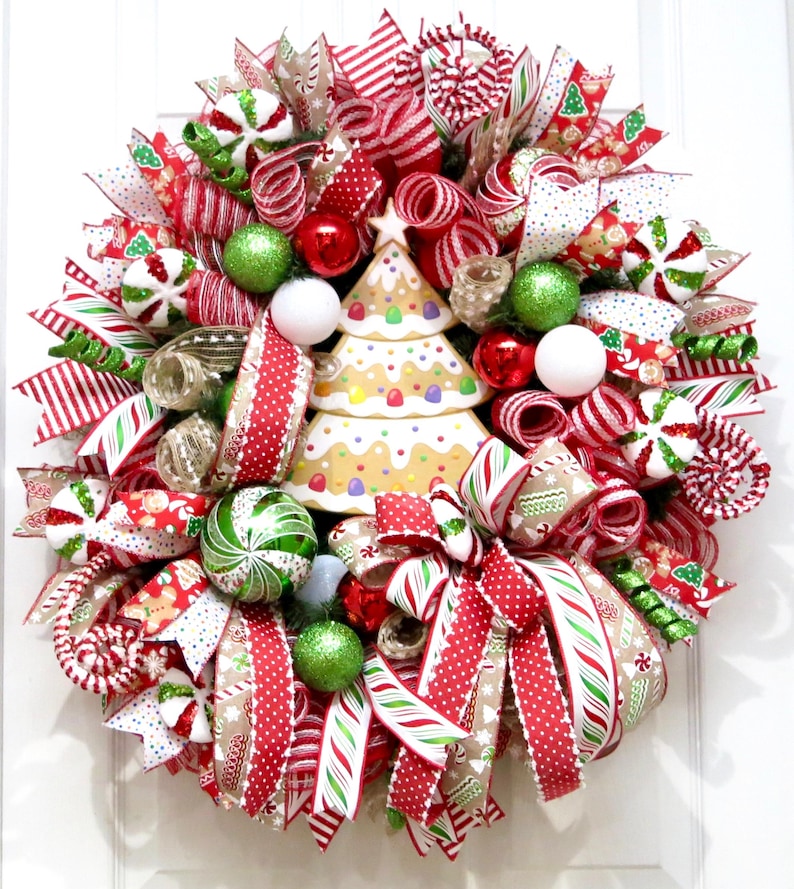 Puede incluir: Una festiva corona navide&ntilde;a con un centro de mesa de &aacute;rbol de pan de jengibre. La corona presenta cintas a rayas rojas y blancas, adornos verdes y rojos y detalles de bastones de caramelo. Un art&iacute;culo decorativo navide&ntilde;o.