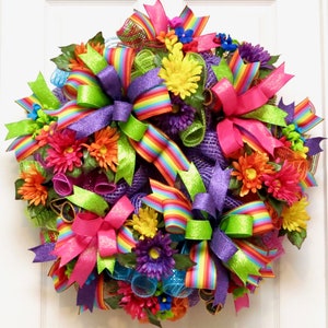 May include: A vibrant, colorful wreath featuring a variety of bright flowers and ribbons. The wreath is adorned with rainbow-striped ribbons, glittery green and purple bows, and an assortment of colorful flowers, creating a cheerful and festive display.