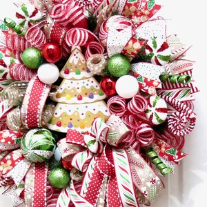Puede incluir: Una corona navide&ntilde;a festiva con un centro de mesa de &aacute;rbol de pan de jengibre. La corona presenta cintas a rayas rojas y blancas, adornos verdes y rojos y detalles de bastones de caramelo. Una alegre decoraci&oacute;n navide&ntilde;a.