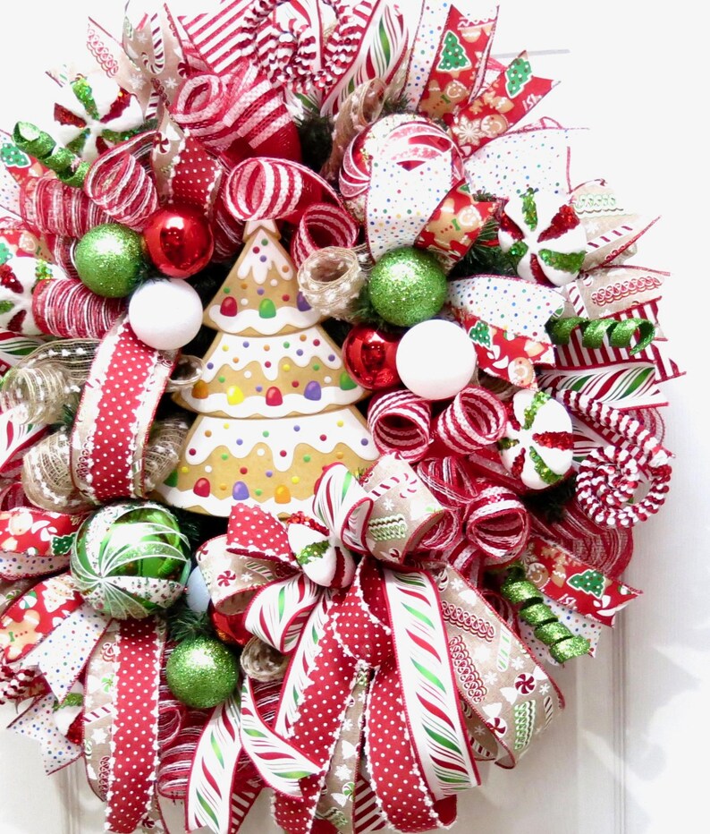 Puede incluir: Una corona navide&ntilde;a festiva con un centro de mesa de &aacute;rbol de pan de jengibre. La corona presenta cintas a rayas rojas y blancas, adornos verdes y rojos y detalles de bastones de caramelo. Una alegre decoraci&oacute;n navide&ntilde;a.