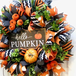 Puede incluir: Una corona oto&ntilde;al festiva con cintas naranjas, negras y blancas, calabazas artificiales y follaje. La corona presenta un letrero que dice "hello PUMPKIN". El dise&ntilde;o incluye lunares, rayas y zigzags, creando una decoraci&oacute;n oto&ntilde;al alegre.