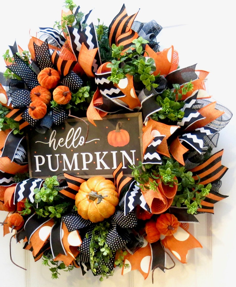 Puede incluir: Una corona oto&ntilde;al festiva con cintas naranjas, negras y blancas, calabazas artificiales y follaje. La corona presenta un letrero que dice "hello PUMPKIN". El dise&ntilde;o incluye lunares, rayas y zigzags, creando una decoraci&oacute;n oto&ntilde;al alegre.