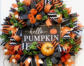 May include: A festive fall wreath with orange, black, and white ribbons, faux pumpkins, and greenery. A small chalkboard sign in the center reads "hello PUMPKIN". The wreath is a circular decoration for a door or wall.