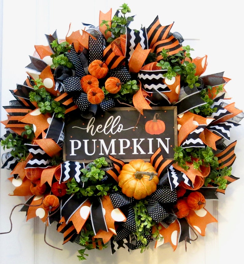 Puede incluir: Una corona oto&ntilde;al festiva con lazos naranjas, negros y blancos, calabazas artificiales y vegetaci&oacute;n. Un peque&ntilde;o letrero de pizarra en el centro dice "hello PUMPKIN". La corona es una decoraci&oacute;n circular para una puerta o pared.
