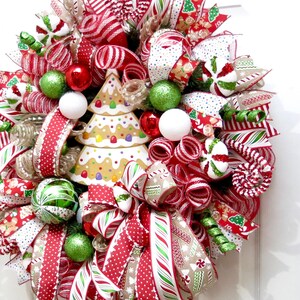 Puede incluir: Una festiva corona navide&ntilde;a con un centro de mesa de &aacute;rbol de pan de jengibre. La corona presenta cintas a rayas rojas y blancas, adornos verdes y rojos y detalles de bastones de caramelo. Un art&iacute;culo decorativo navide&ntilde;o.