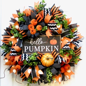 Puede incluir: Una corona oto&ntilde;al redonda con cintas naranjas, negras y blancas, peque&ntilde;as calabazas y follaje verde. Un letrero rectangular en el centro dice "hello PUMPKIN" con una ilustraci&oacute;n de calabaza. Decoraci&oacute;n festiva.