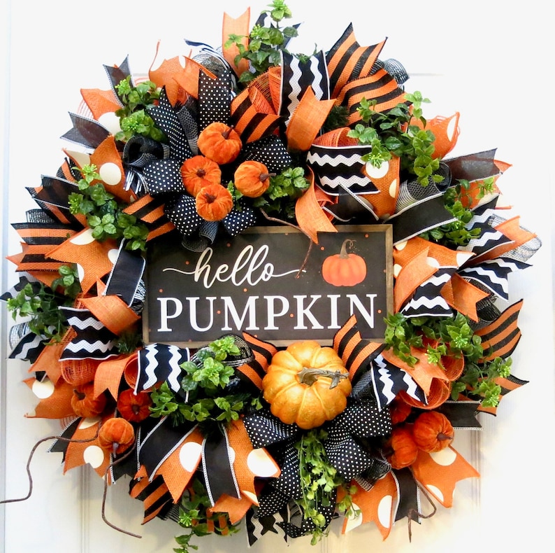 Puede incluir: Una corona oto&ntilde;al redonda con cintas naranjas, negras y blancas, peque&ntilde;as calabazas y follaje verde. Un letrero rectangular en el centro dice "hello PUMPKIN" con una ilustraci&oacute;n de calabaza. Decoraci&oacute;n festiva.