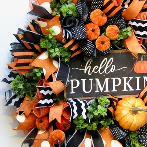 Puede incluir: Una festiva corona de oto&ntilde;o con cintas naranjas, negras y blancas, adornada con peque&ntilde;as calabazas naranjas y vegetaci&oacute;n. Un letrero negro con las palabras "hello PUMPKIN" a&ntilde;ade un toque de temporada a esta corona decorativa.