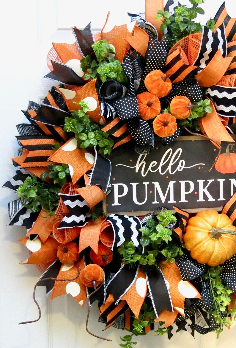 Puede incluir: Una festiva corona de oto&ntilde;o con cintas naranjas, negras y blancas, adornada con peque&ntilde;as calabazas naranjas y vegetaci&oacute;n. Un letrero negro con las palabras "hello PUMPKIN" a&ntilde;ade un toque de temporada a esta corona decorativa.
