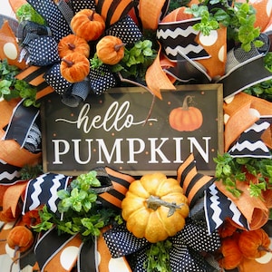 Puede incluir: Una corona oto&ntilde;al festiva con cintas naranjas, negras y blancas, calabazas artificiales y follaje. Un letrero central muestra "hello PUMPKIN" con una imagen de calabaza. Decoraci&oacute;n de temporada.
