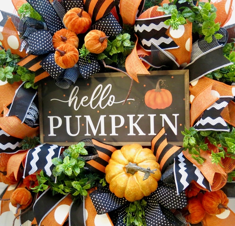 Puede incluir: Una corona oto&ntilde;al festiva con cintas naranjas, negras y blancas, calabazas artificiales y follaje. Un letrero central muestra "hello PUMPKIN" con una imagen de calabaza. Decoraci&oacute;n de temporada.