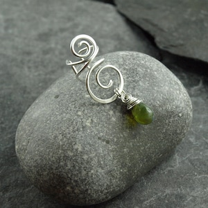 Könnte beinhalten: Ein silberner Ring mit einem Wirbeldesign und einem hängenden grünen Edelstein. Der Ring wird auf einem grauen Stein präsentiert, der die filigrane Drahtarbeit und die lebendige Farbe des Edelsteins hervorhebt. Das Design des Rings ist künstlerisch.