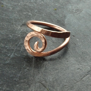 Könnte beinhalten: Kupferfarbener Ring mit gehammerter Textur und Spiraldesign. Der Ring hat ein gehämmertes Band, das sich um den Finger wickelt, und ein dekoratives Spiralelement. Der Ring ist aus Kupfer gefertigt.