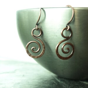 May include: A pair of copper earrings with a spiral design. The earrings are hanging from a metal bowl.