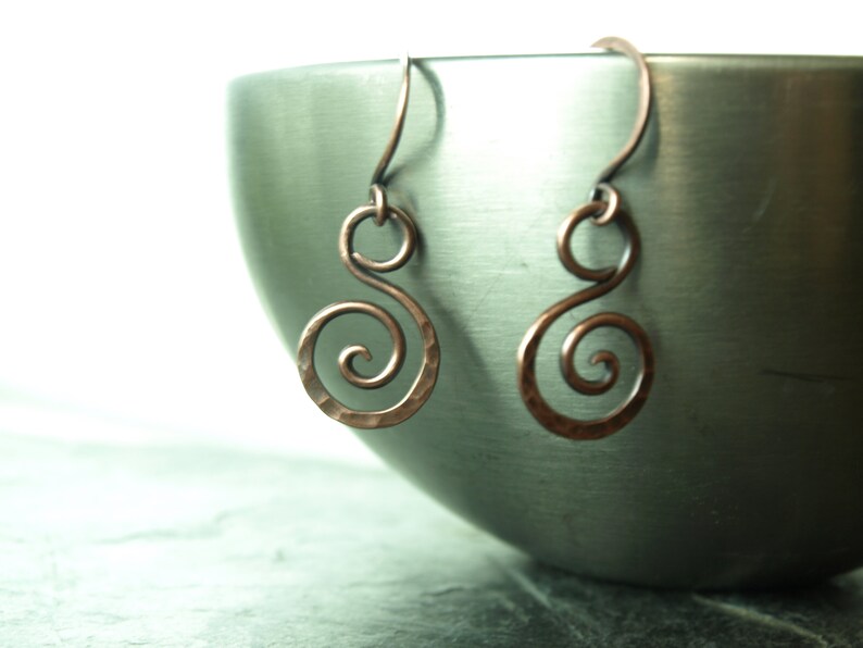 May include: A pair of copper earrings with a spiral design. The earrings are hanging from a metal bowl.