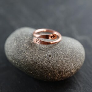 Könnte beinhalten: Ein kupferfarbener Wickelring mit Blattmuster. Der Ring liegt auf einem grauen Stein vor dunklem Hintergrund. Das Design des Rings zeigt ein zartes Blattmuster an jedem Ende und verleiht ihm einen Hauch von naturnaher Eleganz.