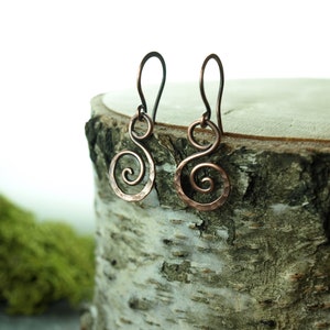 May include: A pair of copper earrings with a hammered finish and a spiral design. The earrings are hanging from a wooden surface.