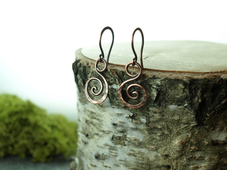 May include: A pair of copper earrings with a hammered finish and a spiral design. The earrings are hanging from a wooden surface.