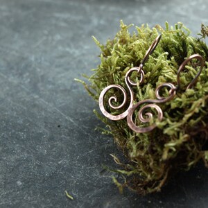 May include: A pair of copper earrings with a swirling design. The earrings are on a bed of green moss.