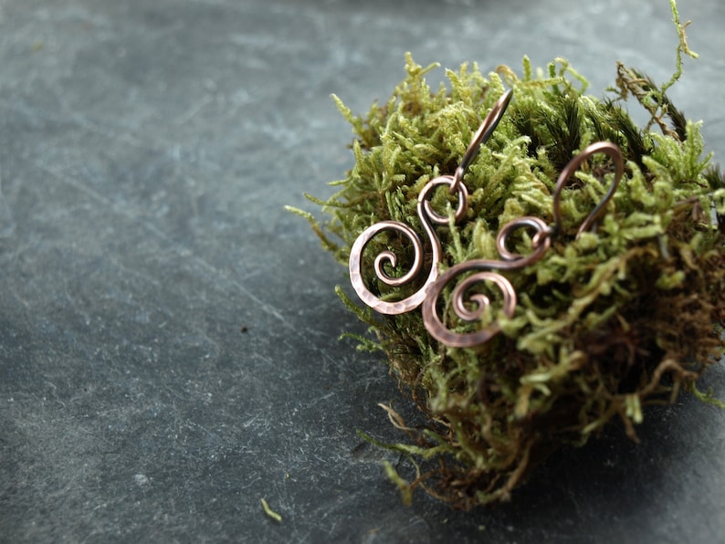May include: A pair of copper earrings with a swirling design. The earrings are on a bed of green moss.