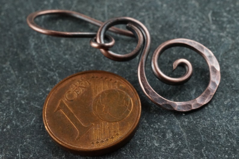 May include: A hammered copper wire earring with a spiral design. The earring is shown next to a 1 cent euro coin for scale.