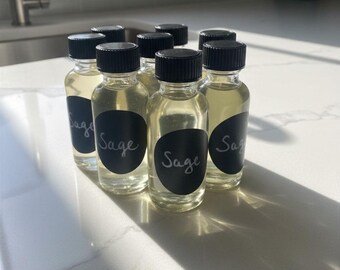 May include: A collection of small, clear glass bottles filled with a light yellow liquid. Each bottle has a black label with the word "Sage" in white script. The bottles are arranged on a white countertop, with sunlight casting shadows.