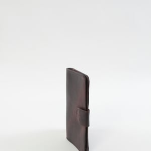 May include: A vertical, dark brown leather wallet with a curved bottom and a flap closure. The wallet is closed and stands upright against a white background. The leather has a textured appearance.