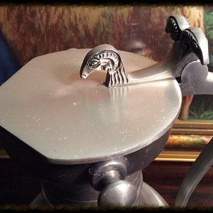 May include: A silver pewter pitcher with a lid featuring a decorative ram's head design. The pitcher has a black handle and a round body.