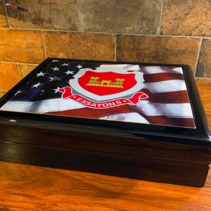 May include: A black, rectangular wooden box with a glossy finish. The lid features an American flag design with a red, white, and blue emblem and the word "ESSAYONS". The box is displayed on a wooden surface.