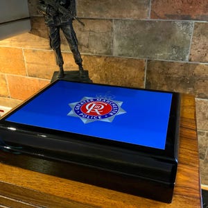 May include: A black wooden box with a blue lid featuring the Greater Manchester Police logo. The logo is a silver star with a red crown and the words "Greater Manchester Police" around it.