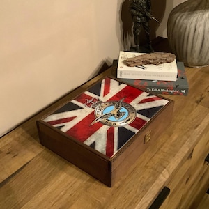 May include: A wooden box with a Union Jack design on the lid. The box features a crest with a bird and the words "PER ARDUA AD ASTRA". The box is on a wooden surface.
