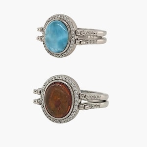 May include: Two silver rings with oval gemstones. One ring features a light blue gemstone, the other a brown gemstone. Both rings have a halo of small, clear stones around the gemstone and additional stones on the band.