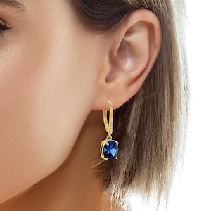 May include: Gold-tone drop earrings featuring a large, oval-cut, deep blue gemstone. The earrings have a lever-back closure and are accented with small, clear crystals along the top portion of the hoop.