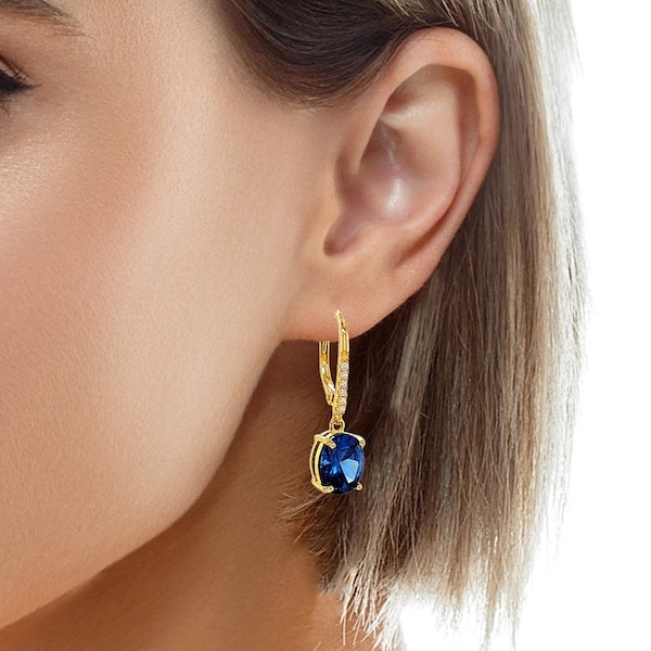 Könnte beinhalten: Goldfarbene Ohrhänger mit einem großen, oval geschliffenen, tiefblauen Edelstein. Die Ohrringe haben einen Klappverschluss und sind mit kleinen, klaren Kristallen verziert.