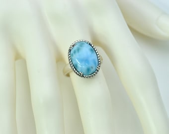 May include: Oval-shaped, light blue gemstone ring with a silver-coloured rope-like border. The ring is set on a white surface. The gemstone has a marbled appearance with shades of blue and white. A piece of jewellery.