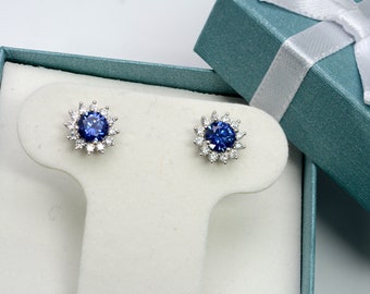 May include: A pair of silver stud earrings with a blue gemstone surrounded by smaller white gemstones. The earrings are displayed on a white stand in a blue gift box.