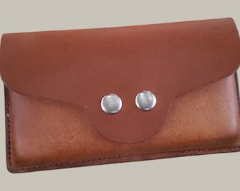 May include: A brown leather wallet with a flap closure secured by two silver snap buttons. The wallet has a rectangular shape and appears to be handmade. The leather has a natural, textured appearance.