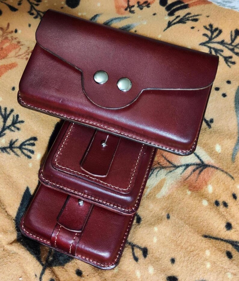 Peut inclure: Portefeuille en cuir bordeaux avec fermeture &agrave; rabat et deux boutons-pression argent&eacute;s. Le portefeuille est empil&eacute; sur deux autres portefeuilles en cuir bordeaux. Les portefeuilles ont un design cousu.