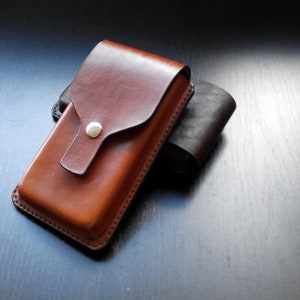 May include: A rectangular brown leather case with a flap closure secured by a single silver-colored metal snap button. The case features visible stitching along the edges and is positioned in front of a second, darker, partially obscured leather case.