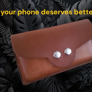Peut inclure: Un &eacute;tui de t&eacute;l&eacute;phone en cuir marron avec une fermeture &agrave; rabat et deux boutons argent&eacute;s. Le texte "your phone deserves better" est affich&eacute; en jaune en haut. L'&eacute;tui est pos&eacute; sur un fond sombre avec des motifs de feuilles.