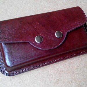 May include: A burgundy leather wallet with a flap closure secured by two silver-colored snaps. The edges are stitched with a contrasting thread, and the leather has a glossy finish.
